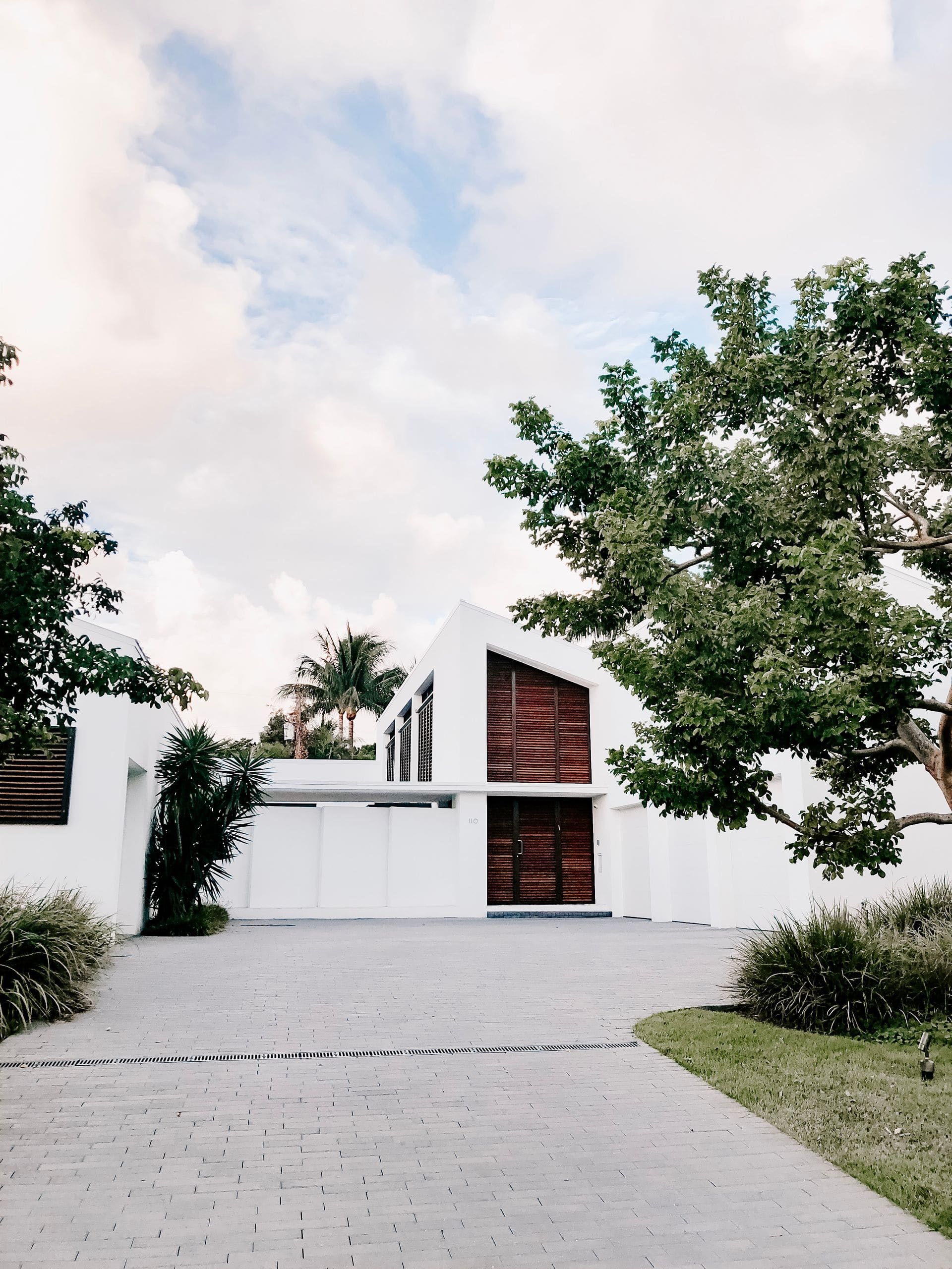 A white two-storey house with timber louvres and a paved forecourt under mature trees