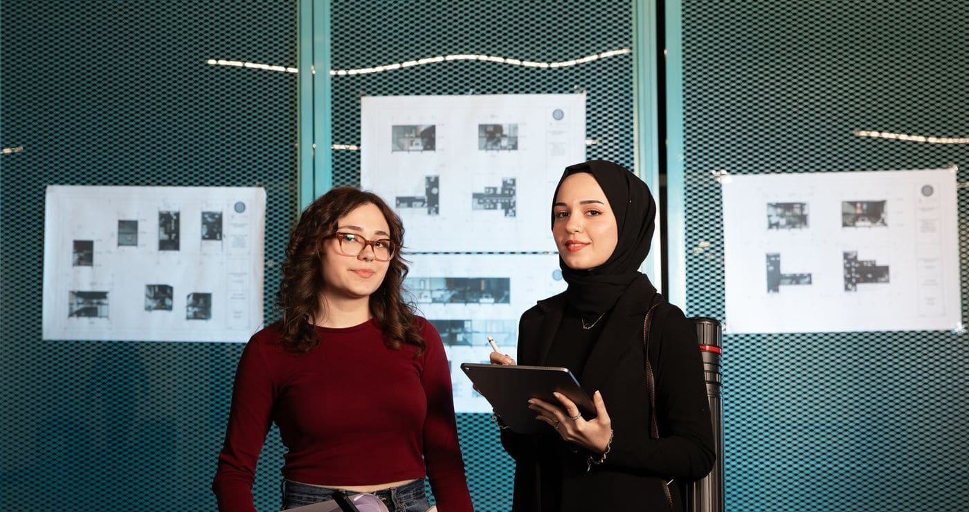 Two architects from the studio holding rolled drawings and a tablet in front of pinned-up plans