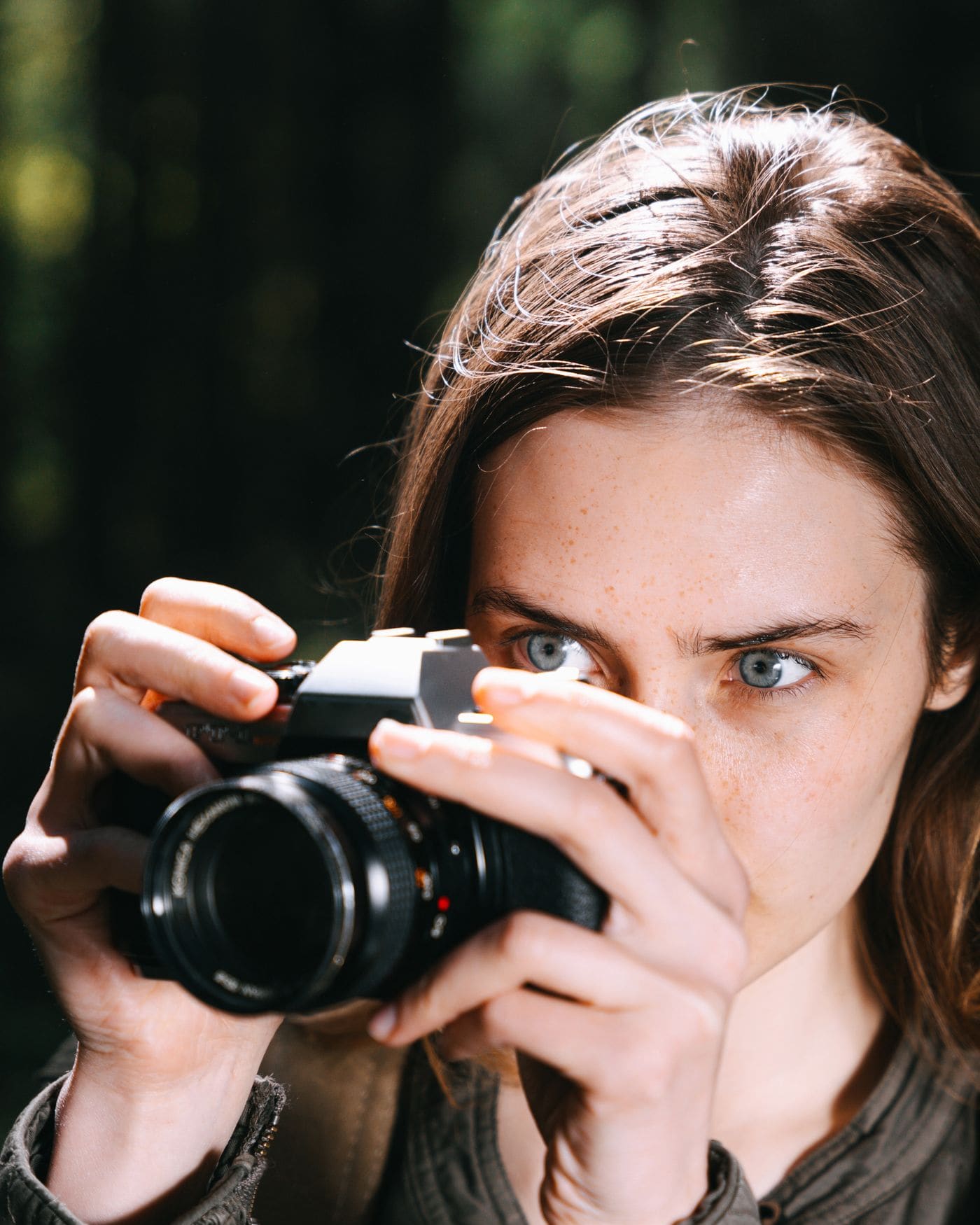 Aisling Byrne holding a film camera to her eye in woodland, looking over the top of it at the camera