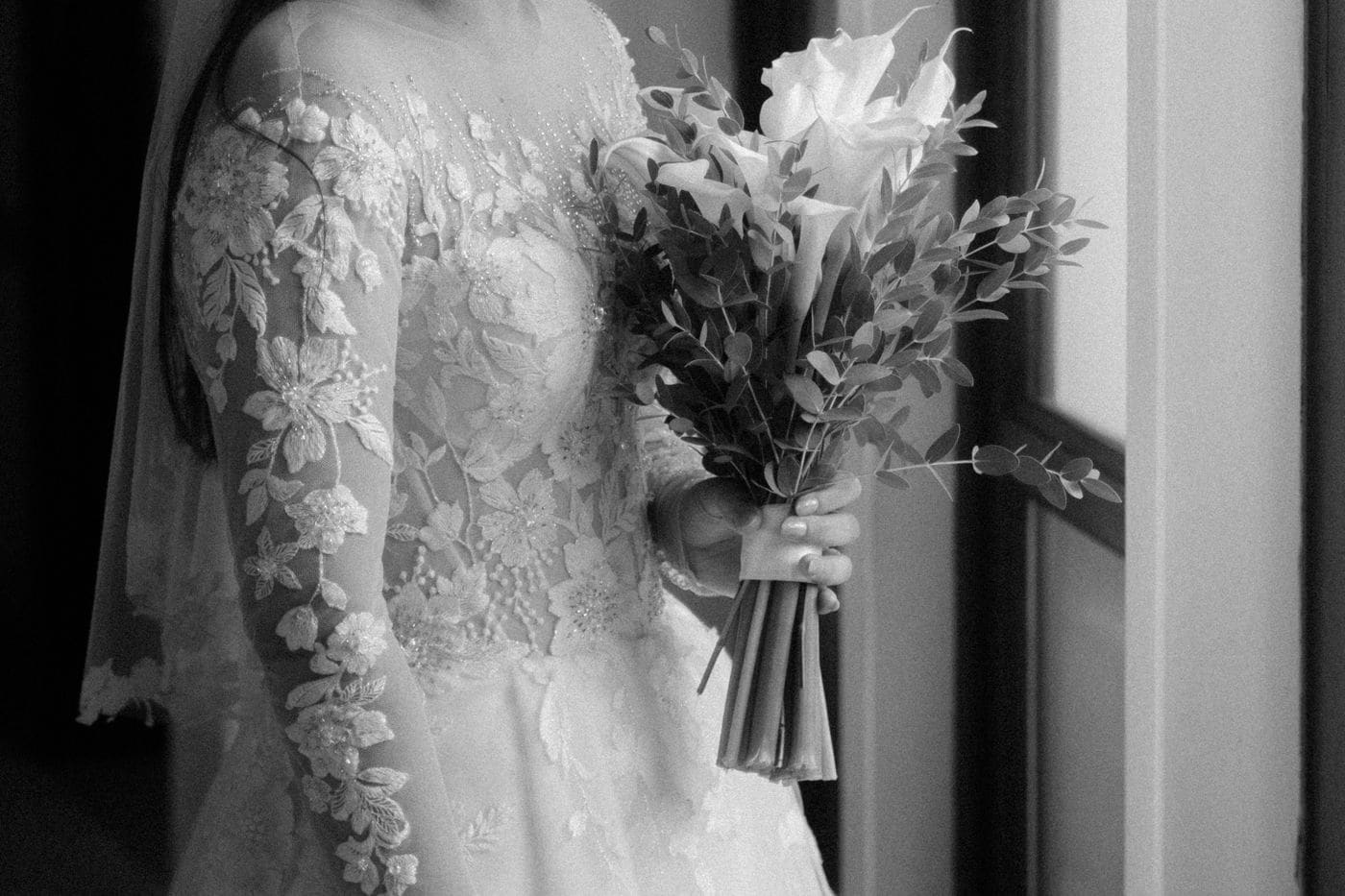 A bride in a lace sleeved gown holding a bouquet of calla lilies beside a window, in black and white