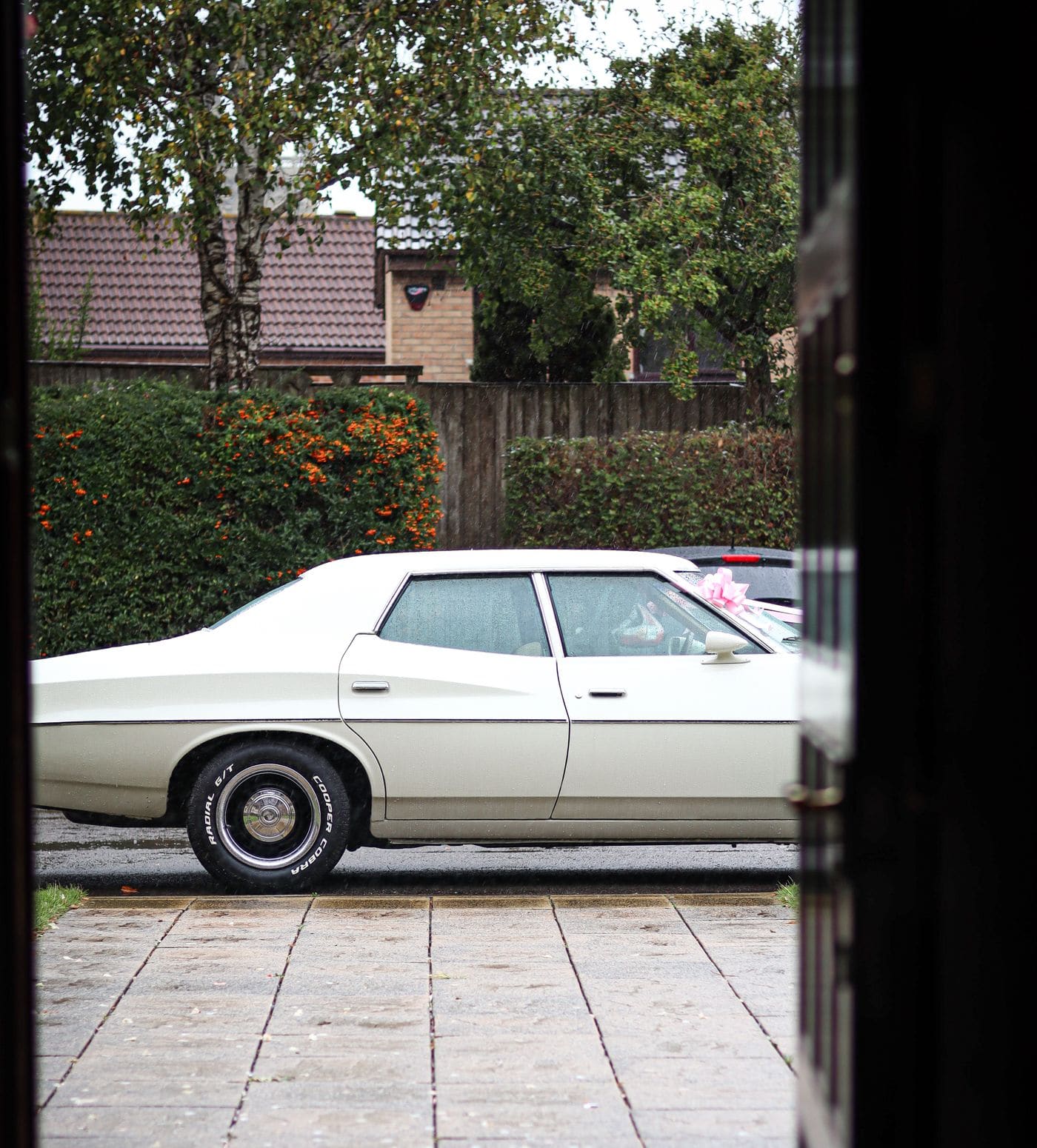 A white nineteen seventies saloon with a ribbon waiting in the rain, seen through the open front door of a house