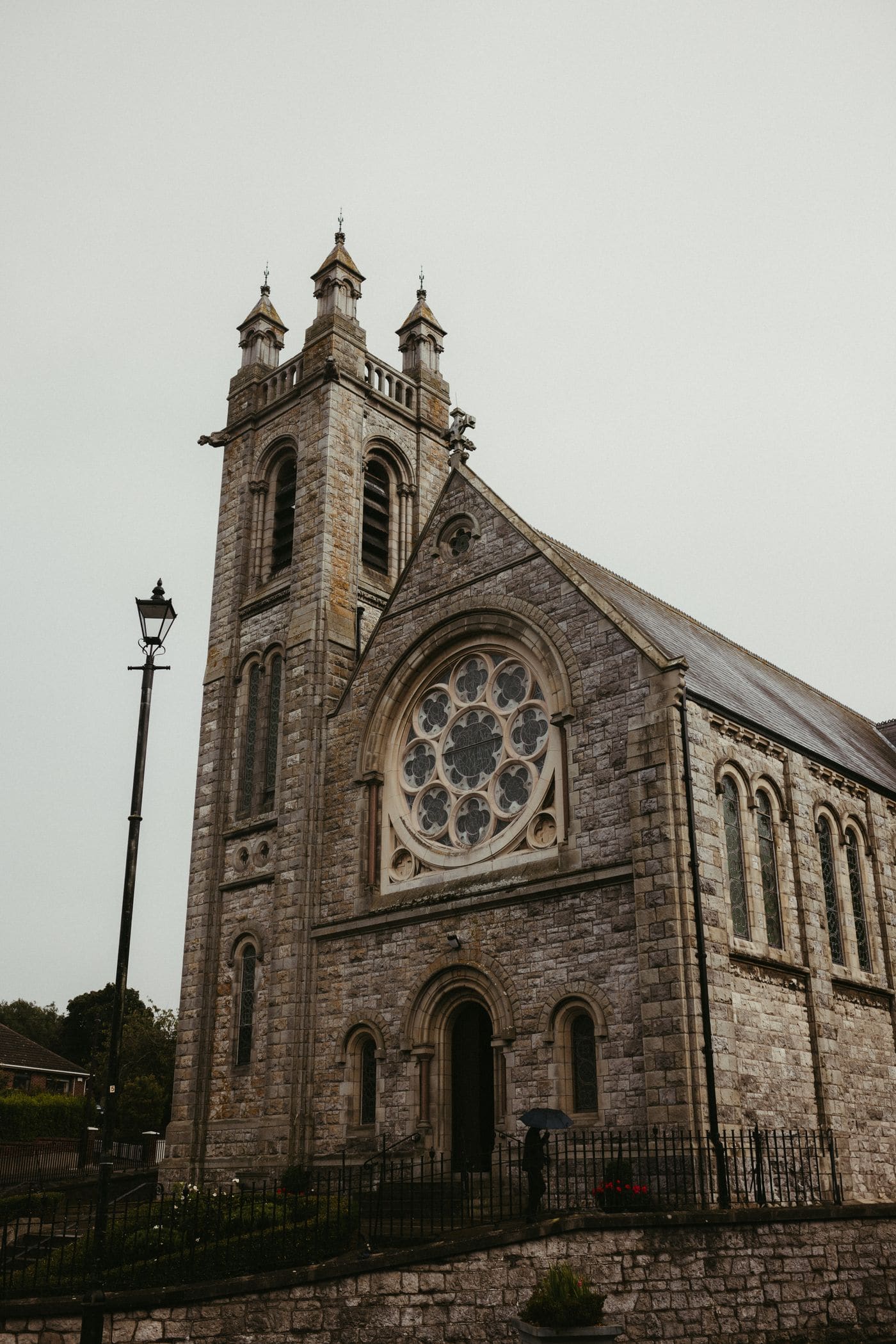 A grey limestone church with a bell tower and rose window under a flat wet sky, a guest sheltering under an umbrella at the door