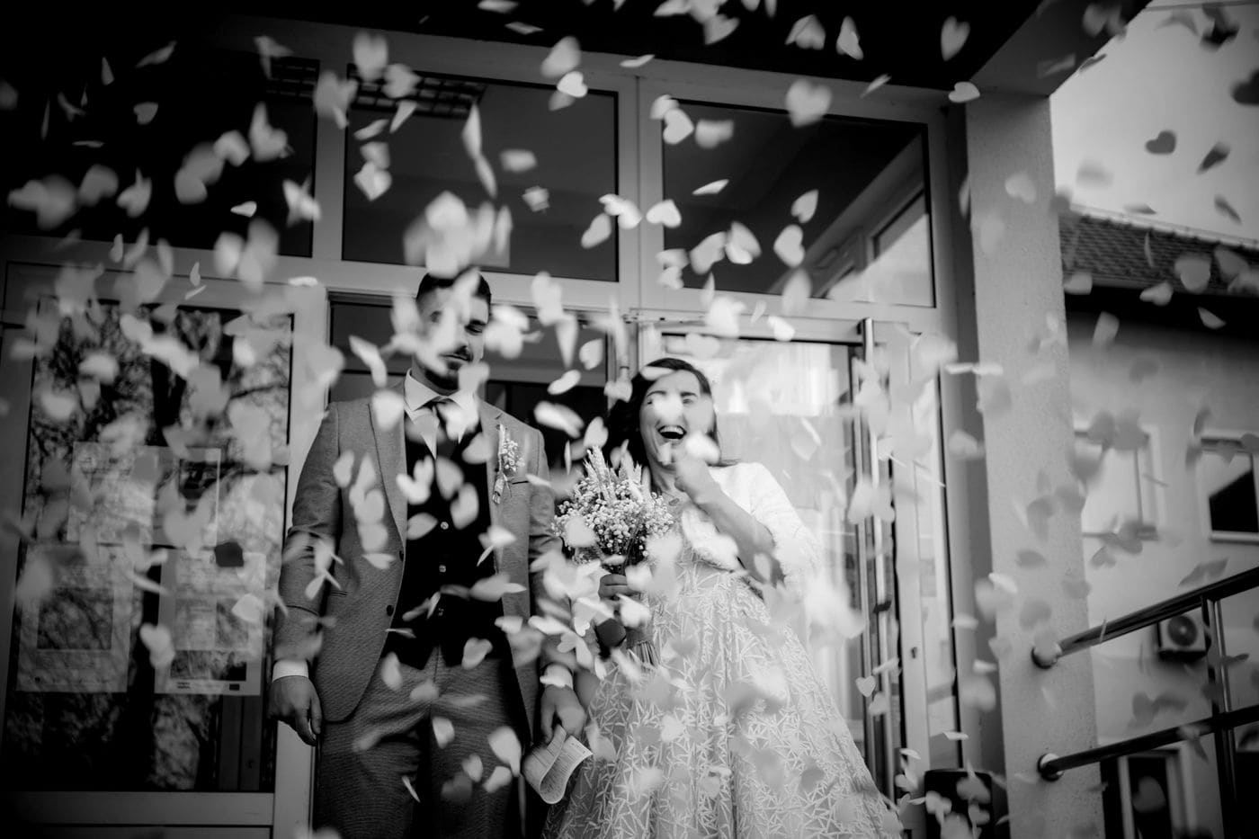 A couple stepping out of a doorway into a shower of paper hearts thrown by guests, in black and white