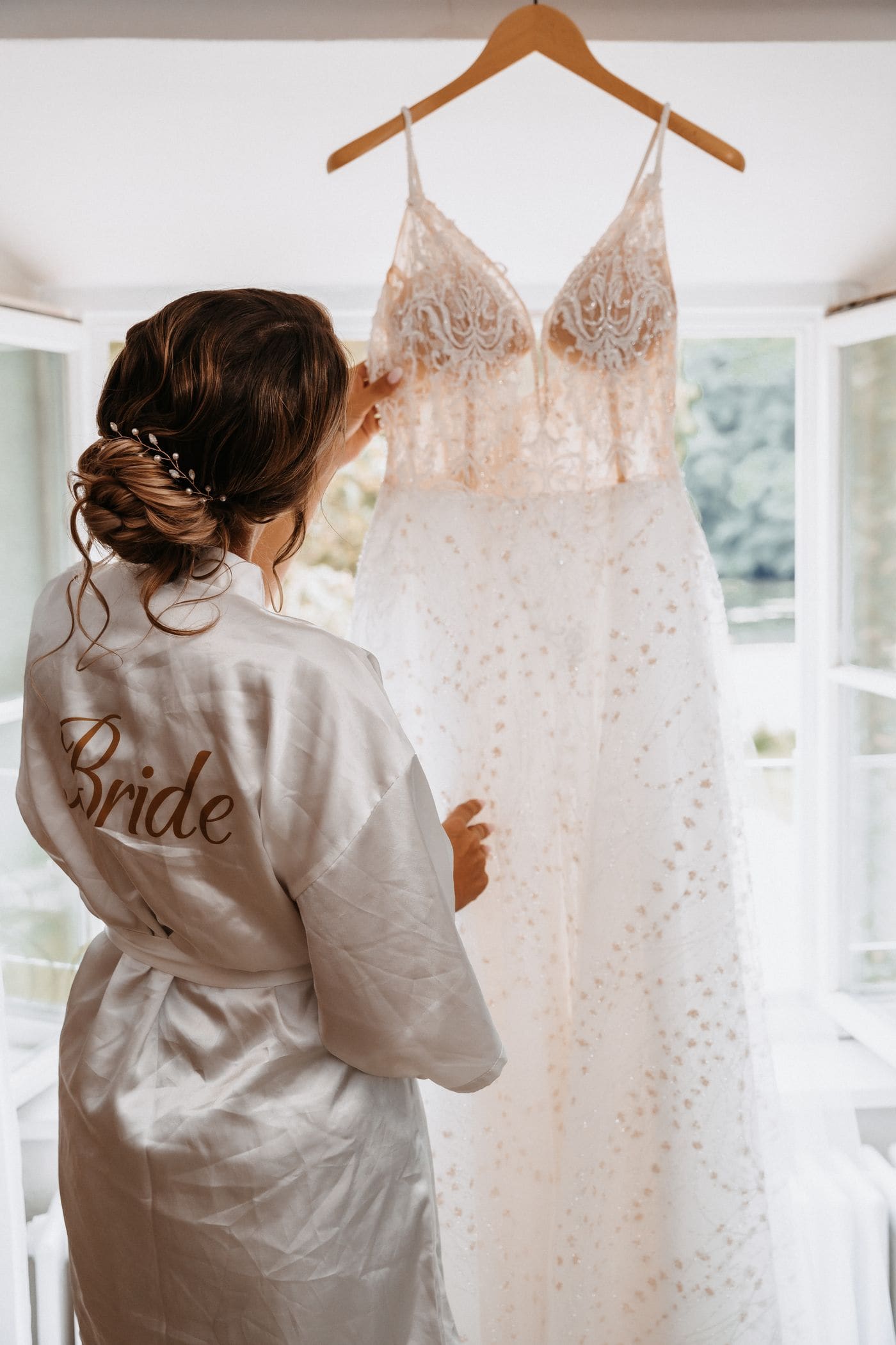 A bride in a robe lifting her wedding dress down from a hanger in front of a bright window