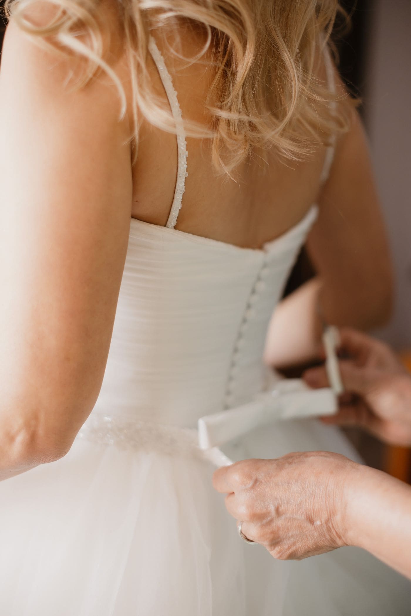 An older woman fastening the back of a wedding gown, close in, lit from a window on the left