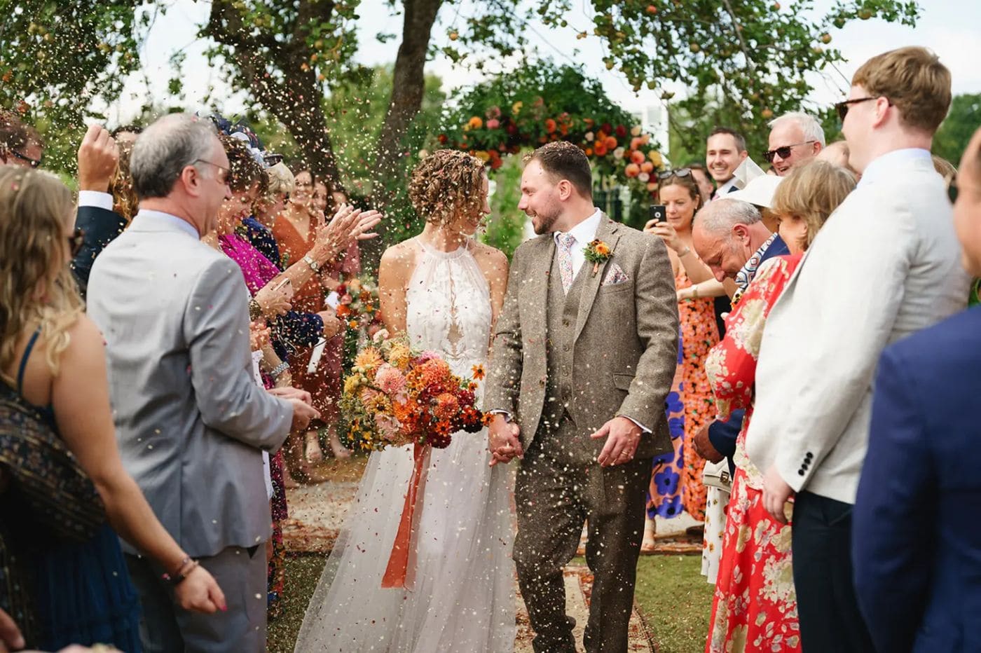Guests throwing confetti over a couple walking out of an outdoor ceremony under an apple tree, the groom in a tweed three piece