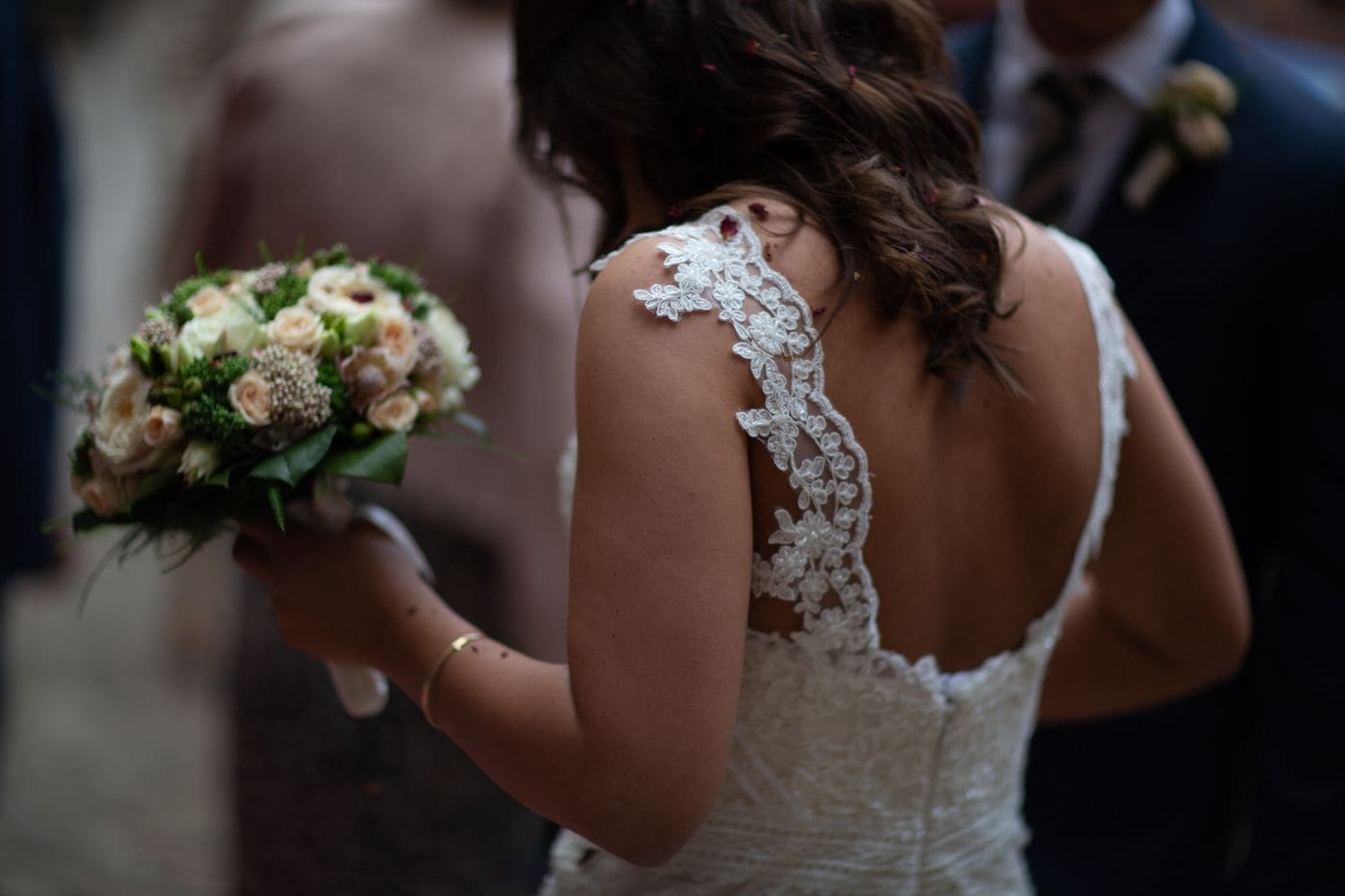 The back of a bride in a lace gown holding a cream bouquet, confetti caught in her hair