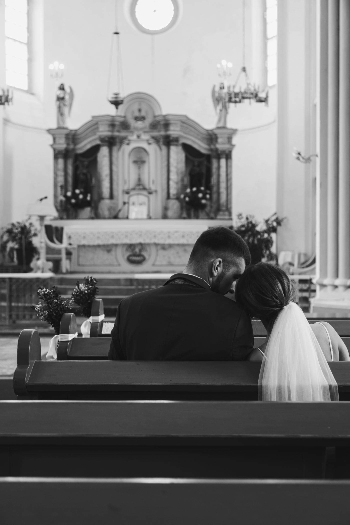 A couple seated together in a church pew seen from behind, her head against his shoulder, the altar beyond