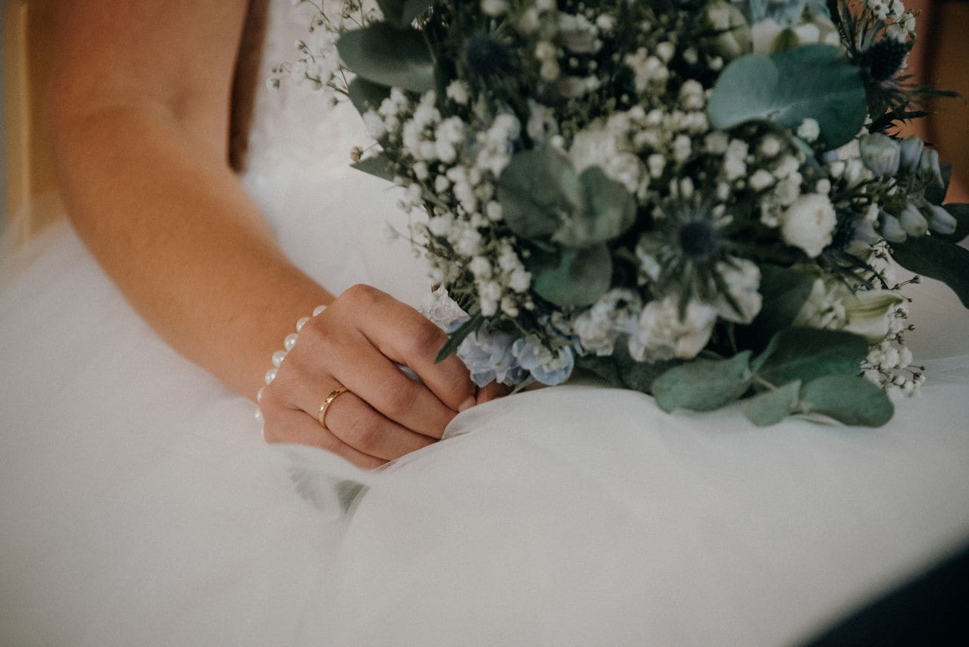 A bride hand resting on a tulle skirt with a plain gold band, beside a bouquet of sea holly, gypsophila and eucalyptus