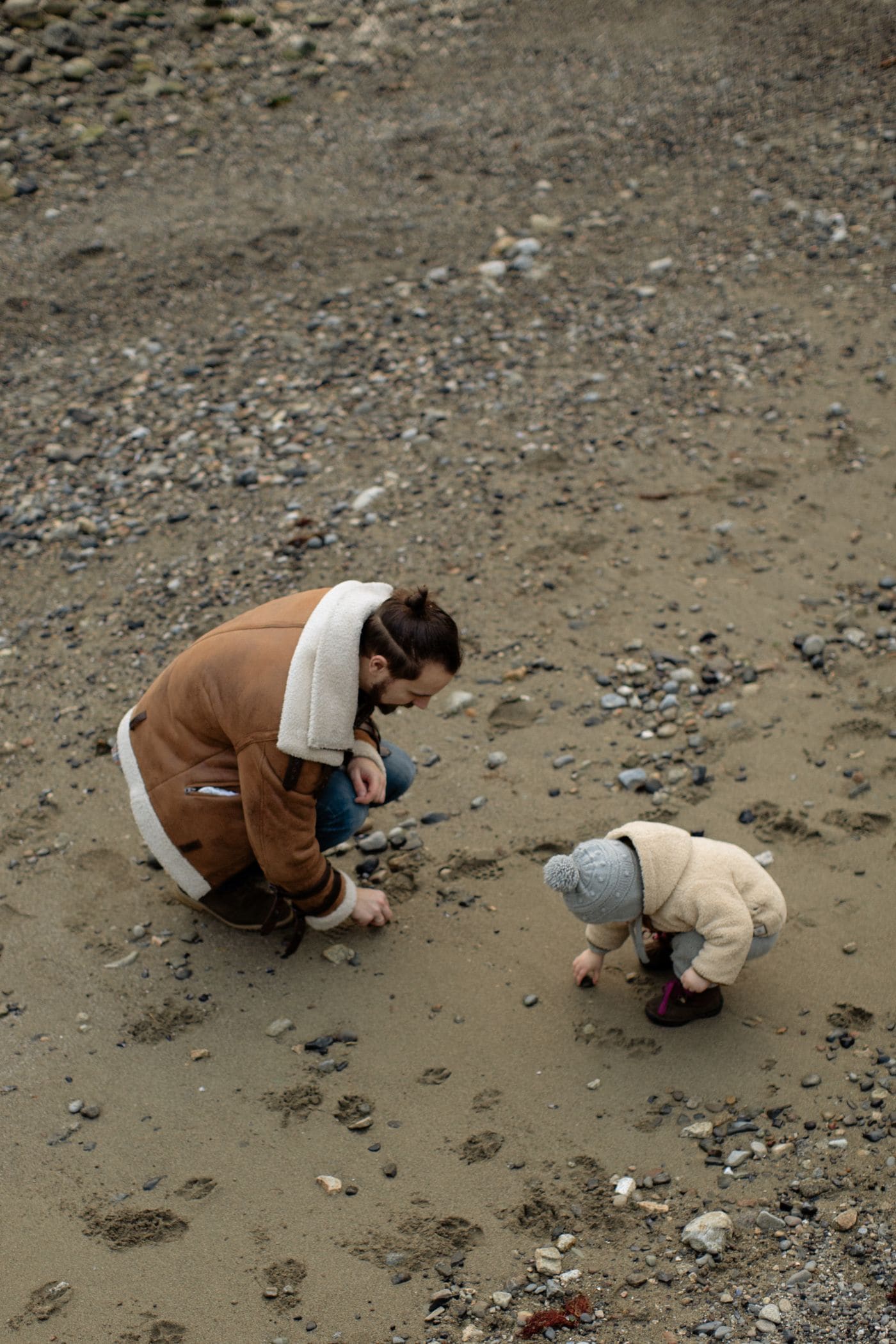 Seen from above, a father crouching on wet sand while a small child in a bobble hat digs beside him