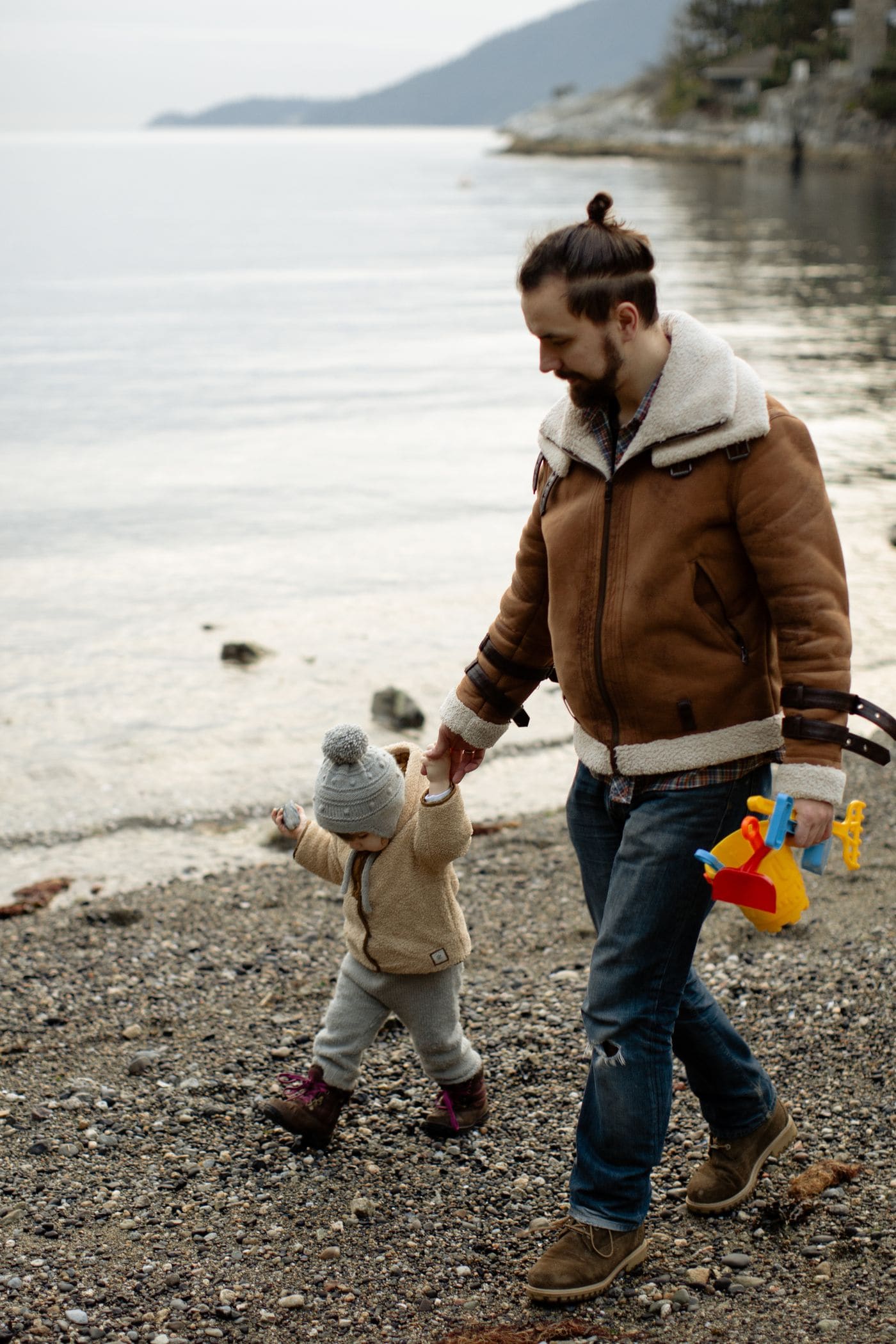 A father in a sheepskin coat walking hand in hand with a toddler along a pebble shore, calm water behind