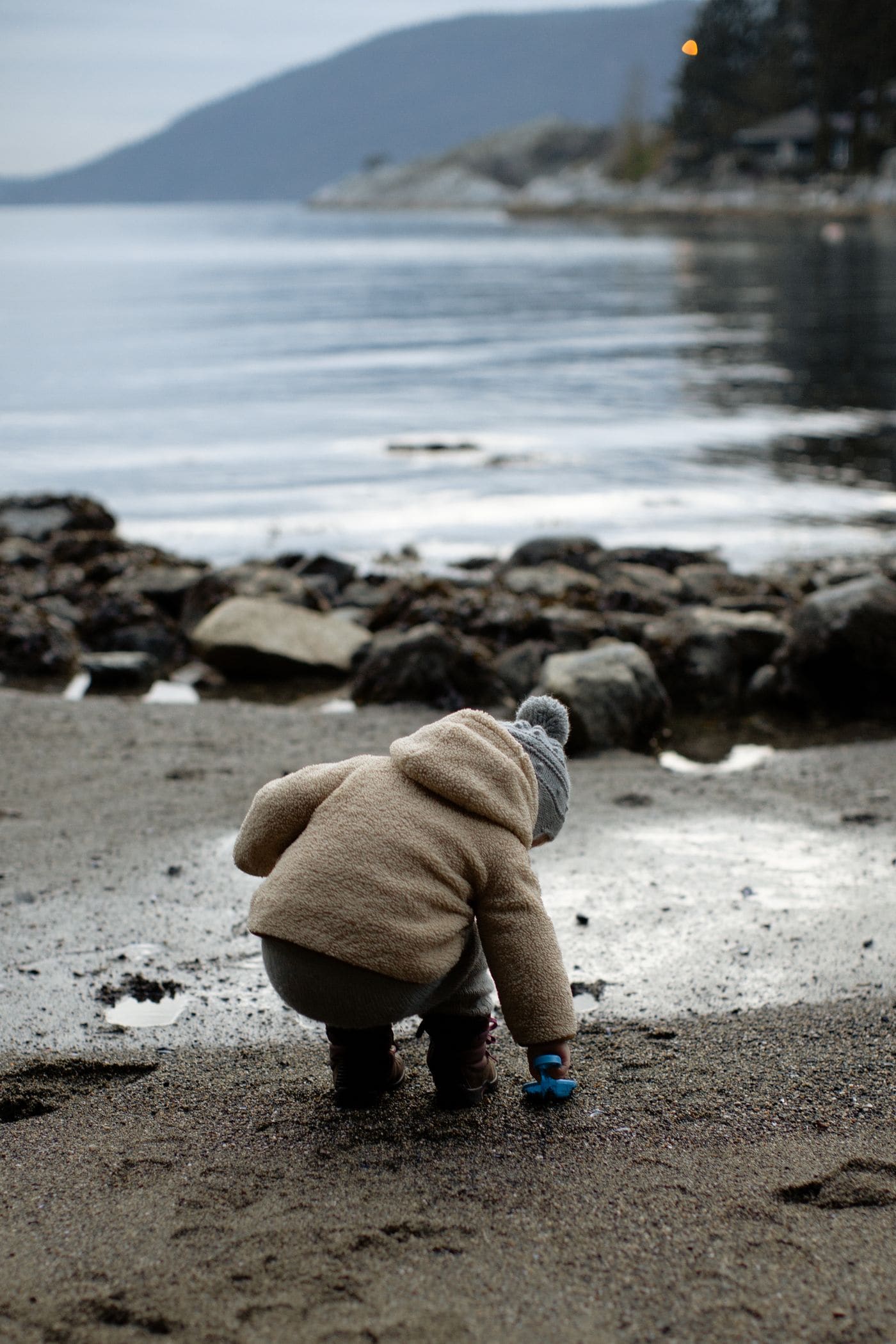 A small child in a fleece coat crouched at the water edge on a pebble beach with hills across the bay behind