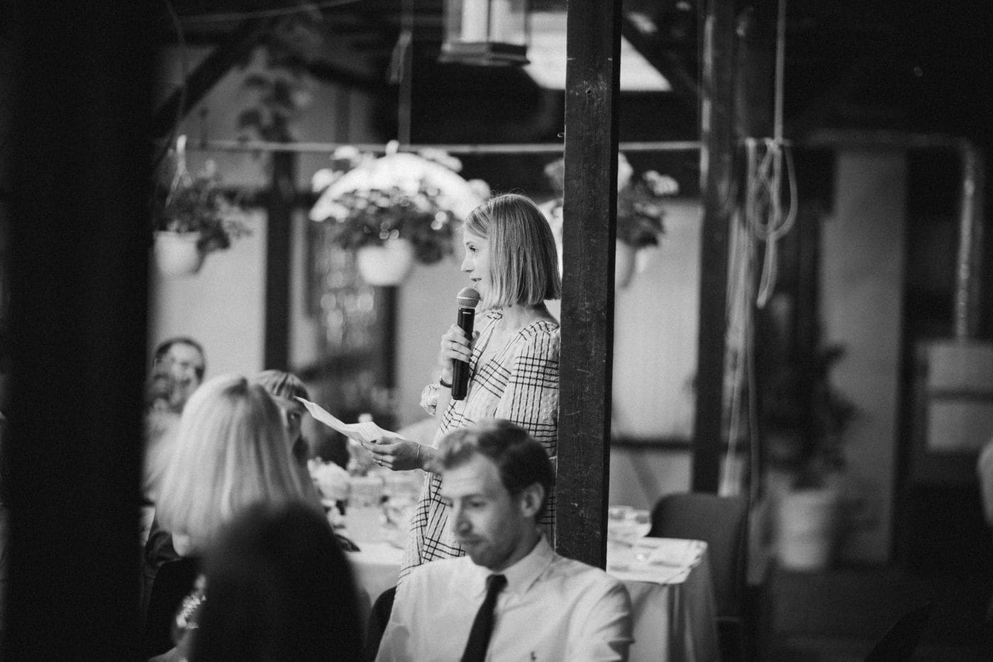 A woman standing to give a speech at a wedding table, reading from a page, guests seated around her