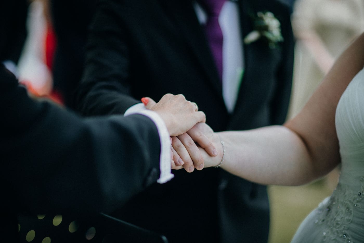 Close view of a couple hands joined during the vows, his dark suit sleeve and her hand over his