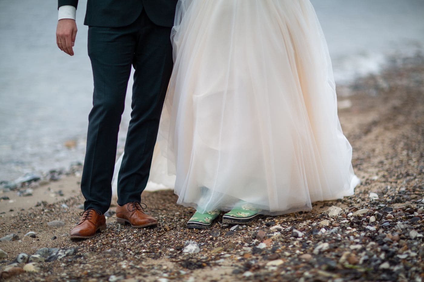 A couple standing on a stony shore photographed from the knees down, the bride in green wellington boots under a tulle skirt